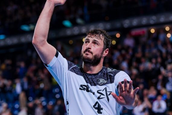 Ein Handballspieler, der ein weißes Trikot mit der Nummer 4 trägt, hebt die rechte Faust und gestikuliert mit der linken Hand. Er steht in einer Halle mit einer verschwommenen Menschenmenge im Hintergrund.