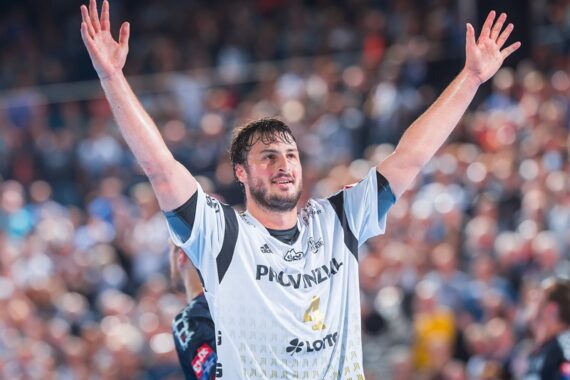 Ein männlicher Handballspieler, der ein weißes Trikot mit der Nummer 4 trägt, hebt beide Arme zum Jubel auf einem Spielfeld.