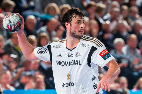 Ein männlicher Handballspieler in einem weißen Trikot bereitet sich darauf vor, den Ball während eines Spiels zu werfen, mit einer unscharfen Zuschauermenge im Hintergrund.