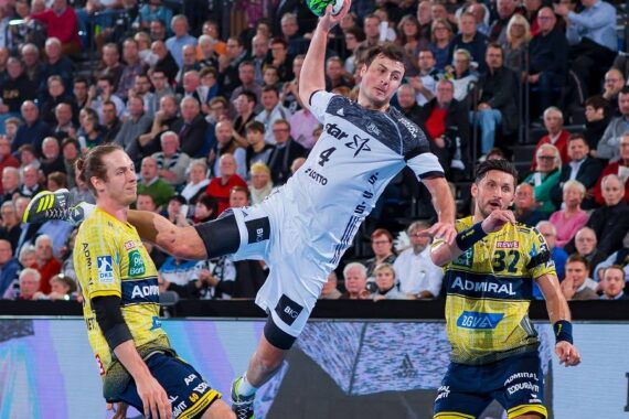 Ein Handballspieler in einem weißen Trikot springt, um den Ball zu werfen, während zwei Verteidiger in gelben und blauen Trikots versuchen, ihn während eines Spiels zu blockieren, während im Hintergrund eine Menschenmenge zusieht.