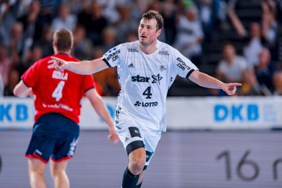 Ein männlicher Handballspieler in einem weißen Trikot mit der Nummer 4 jubelt mit ausgestreckten Armen auf dem Spielfeld, während ein anderer Spieler in einem roten Trikot im Hintergrund davonläuft. Die Tribünen sind mit Zuschauern gefüllt.