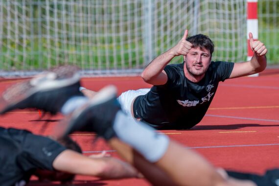 Ein Mann in einem schwarzen Hemd liegt auf einem roten Sportplatz, lächelt und zeigt zwei Daumen nach oben, mit unscharfen Sportlern im Vordergrund und einem Fußballtornetz im Hintergrund.