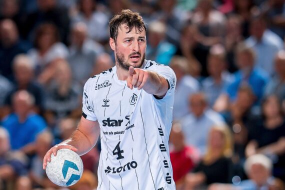 Ein männlicher Handballspieler in einem weißen Trikot hält einen Handball in der linken Hand und zeigt mit der rechten Hand nach vorne auf einem Hallenplatz, mit einer verschwommenen Menschenmenge im Hintergrund.