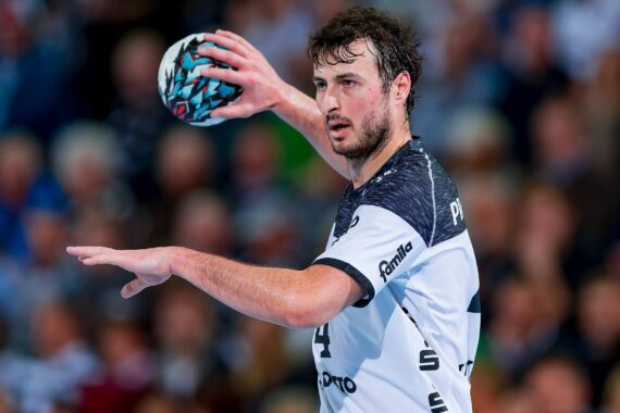 Ein männlicher Handballspieler in einem weiß-schwarzen Trikot hält einen bunten Handball in der Hand und bereitet sich auf den Wurf vor, wobei er einen intensiven Gesichtsausdruck hat. Im Hintergrund ist eine unscharfe Menschenmenge zu sehen.