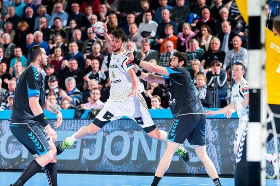 Ein Handballspieler in einem weißen Trikot springt, um den Ball zu werfen, während zwei Verteidiger in dunklen Trikots versuchen, ihn zu blockieren. Das Spiel findet in einer Halle statt, während im Hintergrund eine große Menschenmenge zuschaut.