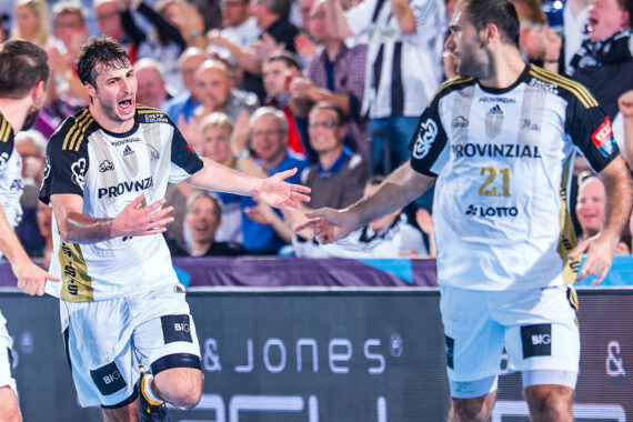 Zwei Handballspieler in weißen Trikots feiern während eines Spiels, umgeben von jubelnden Fans auf der Tribüne. Beide Spieler haben die Arme ausgestreckt und zeigen damit ihre Begeisterung und ihren Enthusiasmus.