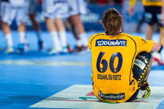 Ein Handballspieler in einem gelben Trikot mit der Nummer 60 und dem Namen Ekdahl Du Rietz sitzt auf dem Spielfeld, von der Kamera abgewandt, während andere Spieler im Hintergrund stehen.