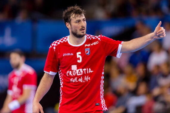 Ein männlicher Handballspieler, der ein rotes Kroatien-Trikot mit der Nummer 5 trägt, zeigt während eines Spiels den Daumen nach oben. Der Hintergrund ist unscharf, Zuschauer und ein weiterer Spieler sind zu sehen.