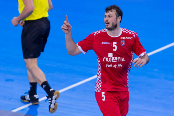Ein männlicher kroatischer Handballspieler in einem roten Trikot mit der Nummer 5 hebt beim Jubeln auf einem blauen Spielfeld den Finger. Ein Beamter in gelben und schwarzen Shorts läuft in der Nähe.
