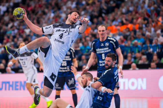 Ein Handballspieler in einem weißen Trikot springt und bereitet sich auf den Wurf vor, während zwei gegnerische Spieler in dunkelblauen Trikots versuchen, ihn während eines Spiels in einer überfüllten Arena zu blockieren.