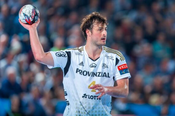 Ein männlicher Handballspieler, der ein weißes Trikot mit der Aufschrift PROVINZIAL und der Nummer 4 trägt, bereitet sich darauf vor, den Ball während eines Spiels zu werfen. Der Hintergrund ist mit Zuschauern unscharf.