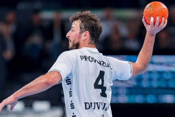 Ein Handballspieler, der ein weißes Trikot mit der Nummer 4 und dem Namen "Duvnjak" trägt, bereitet sich darauf vor, während eines Spiels einen orangefarbenen Handball zu werfen. Der Hintergrund ist unscharf und konzentriert sich auf die Aktion des Spielers.