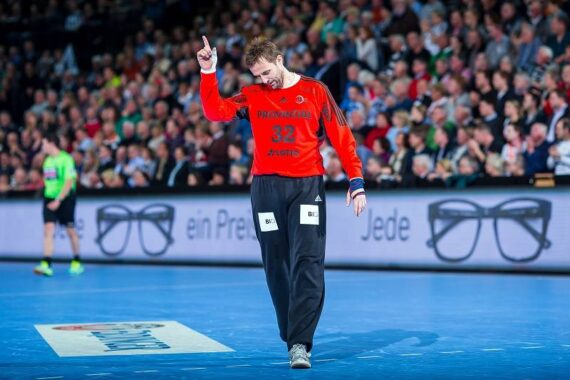 Ein Handballspieler in einem roten Trikot mit der Nummer 32 hebt den Zeigefinger, während er auf das Spielfeld läuft. Im Hintergrund sind eine Zuschauermenge und eine Werbung mit einer Brille zu sehen.