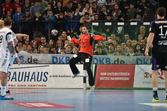 Ein Handballtorwart in einem orangefarbenen Trikot bereitet sich darauf vor, einen Schuss während eines Spiels zu blocken, während gegnerische Spieler in der Nähe sind und eine Menge Zuschauer von der Tribüne aus zusehen.