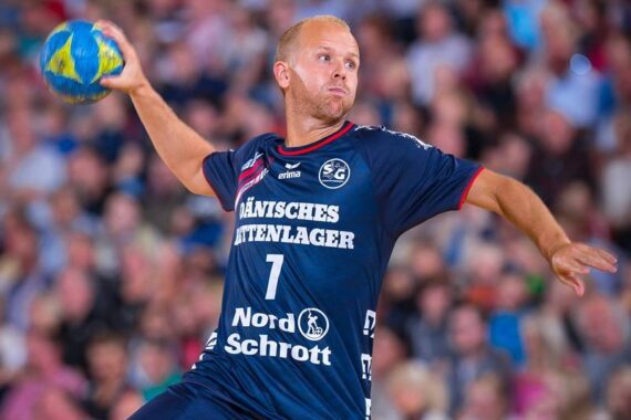 Ein männlicher Handballspieler in einem marineblauen Trikot bereitet sich darauf vor, während eines Spiels einen Ball zu werfen, mit einer unscharfen Menschenmenge im Hintergrund.