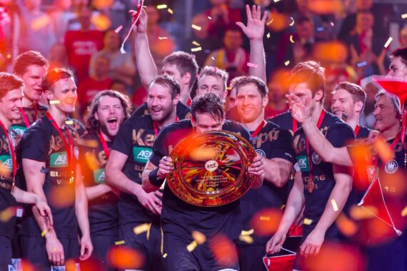 Eine jubelnde Sportmannschaft feiert auf der Bühne, wobei ein Spieler eine große Trophäenplatte in der Hand hält. Konfetti fällt um sie herum, während die Mannschaftskameraden jubeln, lächeln und in festlicher Atmosphäre klatschen.
