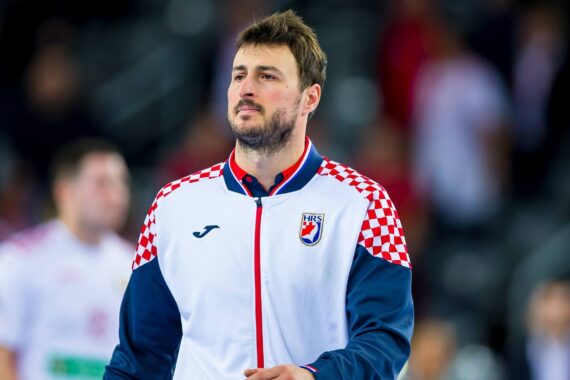 Ein männlicher Sportler, der eine weiß-rot-marineblaue Sportjacke mit einem kroatischen Schachbrettmuster auf den Schultern trägt, steht in einer Halle und schaut nach vorne. Auf der Jacke ist das Logo des kroatischen Handballverbands abgebildet.