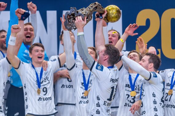 Eine Handballmannschaft in weißen Trikots jubelt, während ein Spieler eine goldene Trophäe über seinen Kopf hebt. Die Mannschaftsmitglieder um ihn herum jubeln und tragen Goldmedaillen.