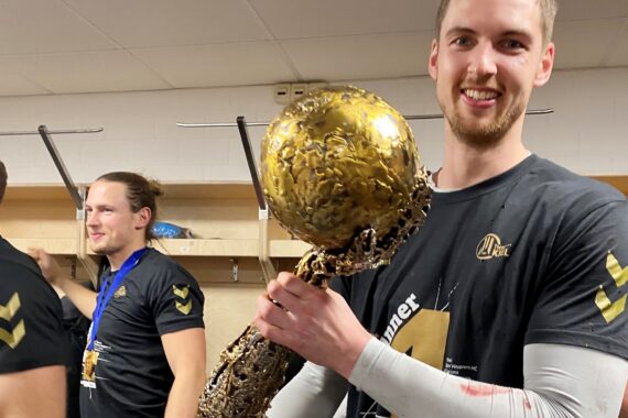 Ein lächelnder Basketballspieler in einem schwarzen Hemd hält in einer Umkleidekabine eine große, goldene Trophäe. Im Hintergrund steht ein weiteres Teammitglied, das ebenfalls eine Medaille und ein Teamshirt trägt.
