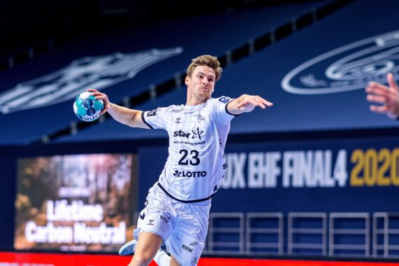 Ein männlicher Handballspieler in einem weißen Trikot bereitet sich in der Luft darauf vor, einen blauen Ball während eines Spiels zu werfen. Im Hintergrund sind das EHF FINAL4 2020 und eine weitgehend leere Arena zu sehen.