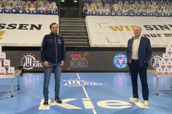 Zwei Männer stehen auf einem Sportplatz mit dem Schriftzug "Kiel" auf dem Boden. Hinter ihnen stapeln sich Kästen mit verschiedenen Logos und Banner mit "THW Kiel" und Vereinslogos. Die Tribüne ist mit Pappfiguren von Fans gefüllt.