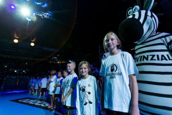 Eine Gruppe von Kindern in weißen Hemden steht in einer Reihe auf dem Boden einer beleuchteten Sportarena, begleitet von einer Person in einem Zebra-Maskottchen-Kostüm. Im Hintergrund sind ein schwach beleuchtetes Publikum und weitere Kinder zu sehen.