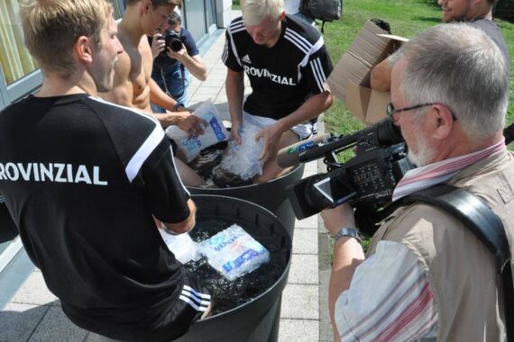 Mehrere junge Männer in Sportuniformen mit der Aufschrift PROVINZIAL sitzen mit ihren Beinen in großen Wannen mit Eiswasser, während andere Eis nachfüllen. Fotografen und ein Kameramann halten die Szene draußen an einem sonnigen Tag fest.