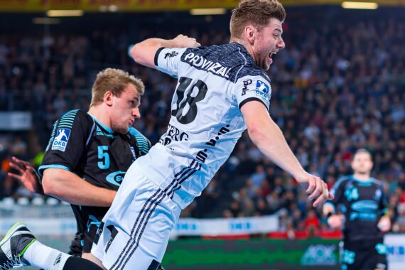 Zwei männliche Handballspieler in Aktion während eines Spiels; der Spieler in Weiß springt vorwärts und hält den Ball, während der Spieler in Schwarz versucht zu verteidigen. Im Hintergrund sieht eine Menschenmenge zu.