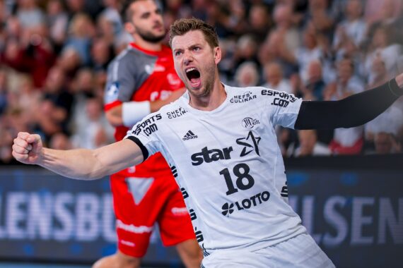 Ein männlicher Handballspieler in einem weißen Trikot mit der Nummer 18 jubelt leidenschaftlich und ballt die Faust. Ein weiterer Spieler in Rot erscheint im Hintergrund. Die Szene spielt sich in einer überfüllten Halle ab.