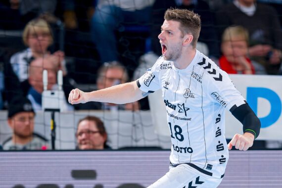 Ein männlicher Handballspieler in einem weißen Trikot mit der Nummer 18 jubelt mit offenem Mund und geballter Faust auf dem Spielfeld. Im Hintergrund sind Zuschauer und ein Tornetz zu sehen.