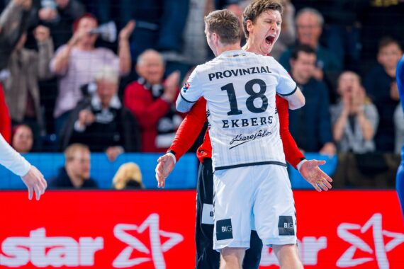 Zwei männliche Handballspieler in weißen Trikots feiern leidenschaftlich auf dem Spielfeld und umarmen sich, während jubelnde Fans im Hintergrund klatschen und Fotos machen. Ein Spieler hat PROVINZIAL 18 EKBERG auf seinem Trikot.