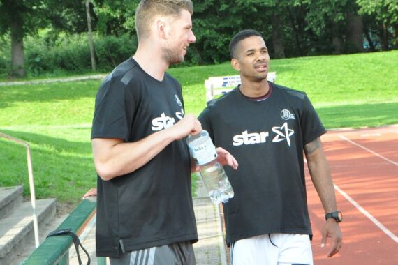 Zwei Männer stehen am Rande einer Laufbahn im Freien, unterhalten sich und lächeln. Beide tragen schwarze Sporthemden mit aufgedruckten Sternen. Einer hält eine Wasserflasche in der Hand. Im Hintergrund sind grünes Gras und Bäume zu sehen.