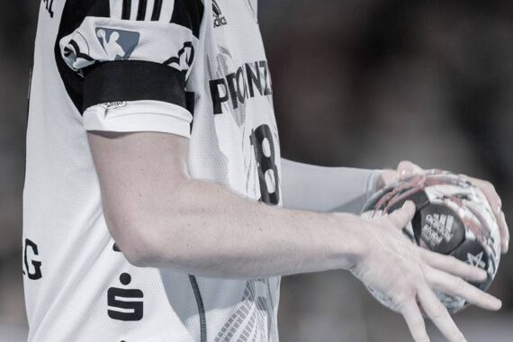 Eine Nahaufnahme eines Handballspielers in einem weißen Trikot, der einen Handball hält, wobei nur der Oberkörper und die Arme zu sehen sind. Das Trikot des Spielers hat schwarze Streifen und sichtbare Sponsorenlogos.