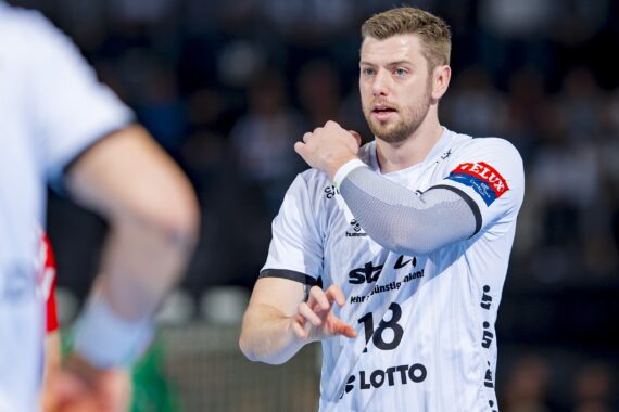 Ein männlicher Handballspieler in einem weißen Trikot mit der Nummer 18 rückt seinen Ärmel auf dem Spielfeld zurecht, wobei der Arm eines anderen Spielers im Vordergrund zu sehen ist. Der Hintergrund ist mit Zuschauern unscharf.