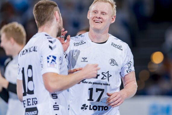Zwei männliche Handballspieler in weißen Trikots mit den Nummern 17 und 18 lächeln und unterhalten sich während eines Spiels. Der Hintergrund ist unscharf, andere Spieler und Zuschauer sind zu sehen.