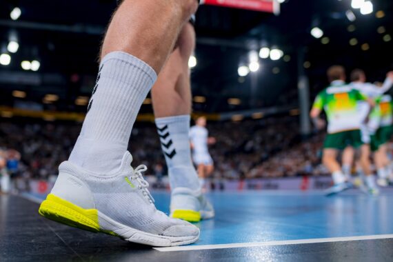 Nahaufnahme des Fußes eines Handballspielers mit weiß-gelbem Schuh und weißer Socke auf dem Spielfeldrand, mit einem unscharfen Hintergrund anderer Spieler und einer überfüllten Arena.