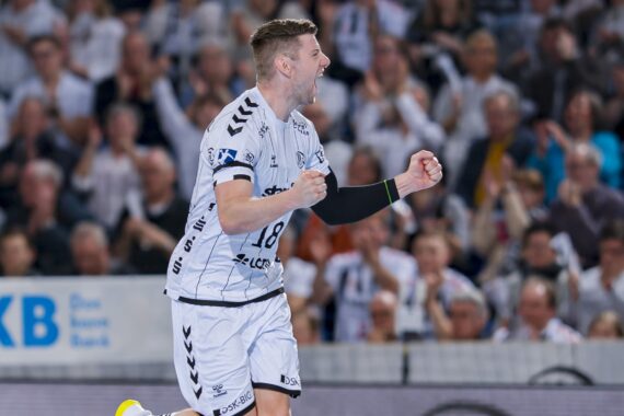 Ein männlicher Handballspieler in einem weißen Trikot feiert leidenschaftlich auf dem Spielfeld und ballt die Fäuste, während im Hintergrund eine unscharfe Menge von jubelnden Zuschauern zu sehen ist.