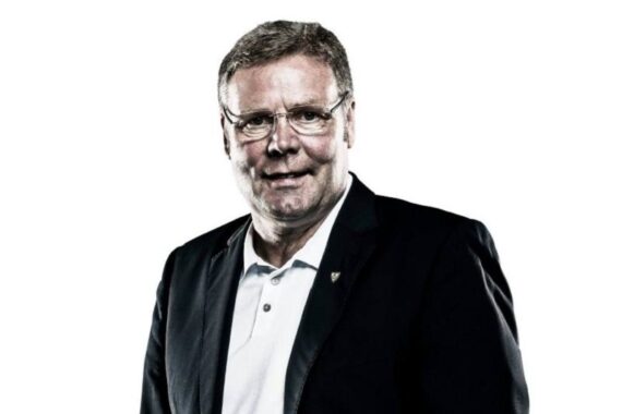 Ein Mann mittleren Alters mit kurzen hellbraunen Haaren und Brille, der ein weißes Hemd mit Kragen unter einem dunklen Blazer trägt, posiert vor einem schlichten weißen Hintergrund.