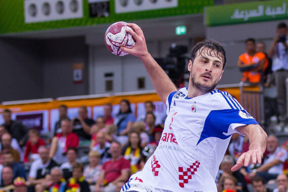 Ein männlicher Handballspieler in einem weiß-blauen Trikot springt in die Luft, hält einen Ball und bereitet sich darauf vor, ihn zu werfen. Die Zuschauer schauen von der Tribüne im Hintergrund zu.