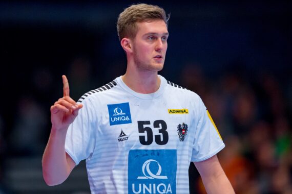 Ein männlicher Handballspieler in einem weißen Trikot mit der Nummer 53 hebt den rechten Zeigefinger. Er scheint konzentriert zu sein, während die Zuschauer im Hintergrund verschwommen sind.