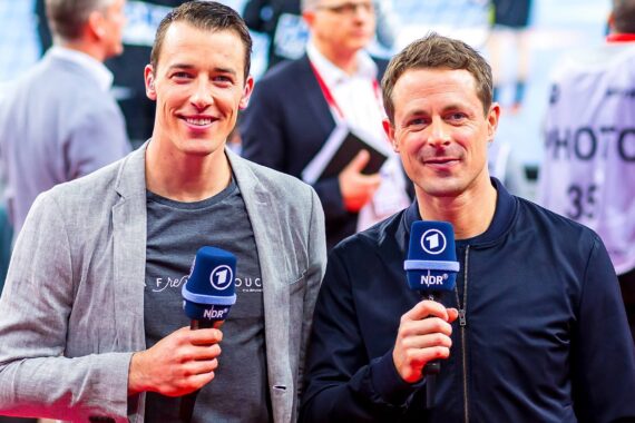 Zwei Männer stehen nebeneinander, halten Mikrofone mit NDR- und 1-Logo und lächeln in die Kamera. Sie befinden sich in einer hellen Halle, möglicherweise bei einer Sportveranstaltung, mit unscharfen Menschen im Hintergrund.