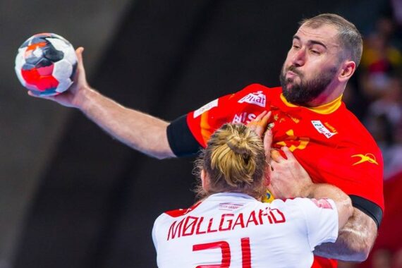 Ein männlicher Handballspieler in einem roten Trikot versucht zu schießen, während er von einem Gegner in einem weißen Trikot mit der Aufschrift MØLLGAARD 21 auf dem Rücken während eines Spiels eng verteidigt wird.