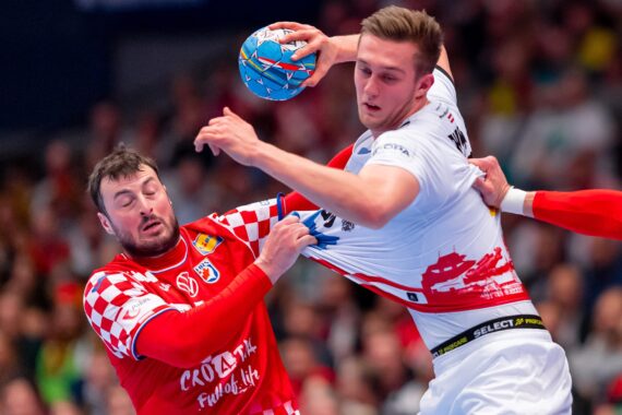 Zwei männliche Handballspieler liefern sich einen erbitterten Wettkampf; der Spieler in Rot greift nach dem Trikot des weiß gekleideten Spielers, während er nach dem Ball greift, und beide zeigen während des Spiels großen Einsatz und Entschlossenheit.