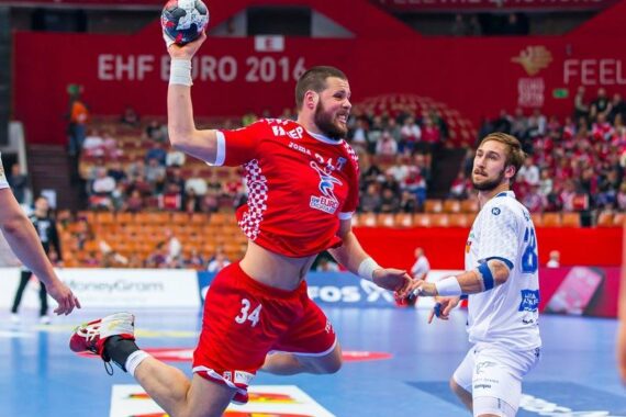 Ein Handballspieler in einem roten Trikot springt in der Luft, um den Ball zu werfen, während ein Verteidiger in einem weißen Trikot während eines Spiels bei der EHF Euro 2016 aufmerksam zusieht. Die Zuschauer füllen die Tribünen im Hintergrund.