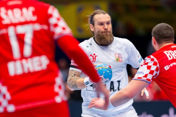 Ein bärtiger männlicher Handballspieler in einem weißen Trikot hält den Ball und steht während eines Spiels zwei Gegnern in rot karierten Trikots gegenüber.