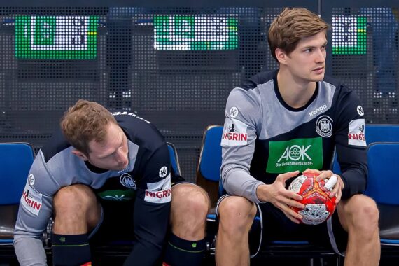 Zwei männliche Handballspieler in schwarz-grauen Uniformen sitzen auf einer Bank; einer bindet sich die Schnürsenkel, während der andere einen Ball hält und zur Seite schaut. Sie scheinen darauf zu warten, mit dem Spiel beginnen zu können.