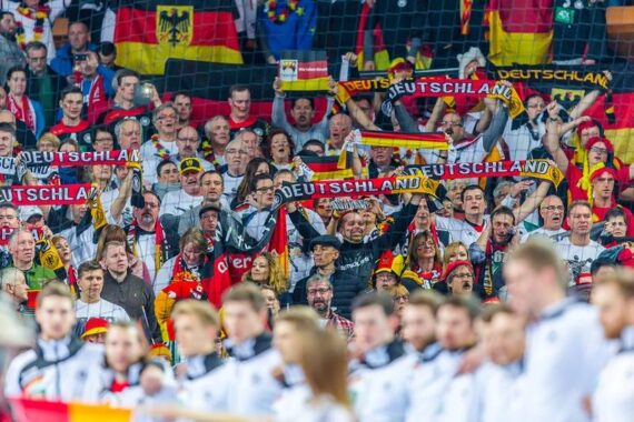 Eine große, enthusiastische Menge deutscher Sportfans, viele mit Deutschland-Schals und deutschen Fahnen, jubelt von der Tribüne aus. Die Fans tragen Nationalfarben und Trikots und schaffen eine lebendige, energiegeladene Atmosphäre.