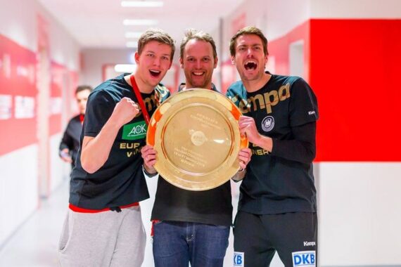 Drei Männer in Sportkleidung posieren fröhlich in einem Korridor und halten eine große goldene Meisterschaftsplakette mit Gravuren. Sie wirken aufgeregt und feierlich, wahrscheinlich nach dem Gewinn eines Sportereignisses.