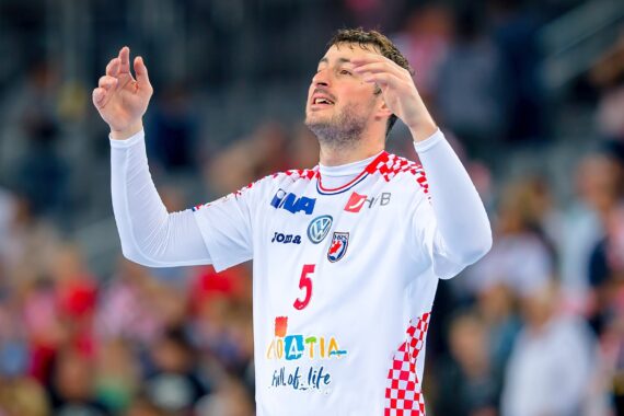 Ein männlicher Handballspieler in einem weiß-roten Trikot mit der Nummer 5 hebt während eines Spiels die Arme, während im Hintergrund eine unscharfe Menschenmenge zu sehen ist.