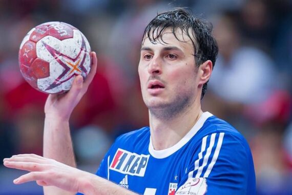 Ein männlicher Handballspieler in einem blauen Trikot bereitet sich auf den Wurf eines rot-weißen Handballs während eines Spiels vor. Er scheint konzentriert zu sein, während im Hintergrund ein unscharfes Publikum zu sehen ist.
